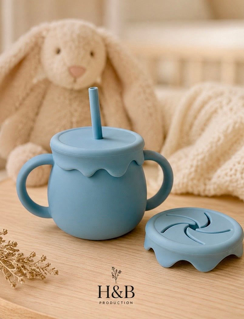 A light blue baby tumbler with integrated handles, a matching lid, and straw, displayed on a wooden surface with a plush bunny toy and soft blanket in the background. The brand logo 'H&B PRODUCTION' is visible at the bottom.