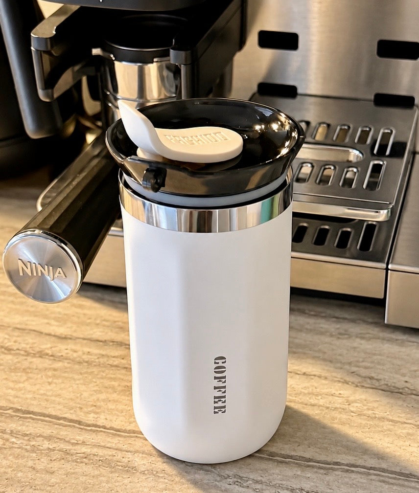 A white insulated coffee cup with a black lid and small white spoon, standing on a countertop in front of a coffee machine. The cup has the word COFFEE printed vertically on the front, and a visible NINJA logo appears on the coffee machine behind it.