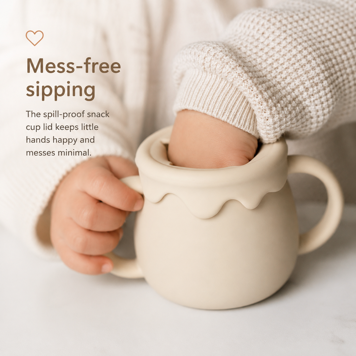 A child's hand and arm in a cream knit sweater insert into a spill-proof cream ceramic snack cup with a handle, on a light surface with promotional text on the left.