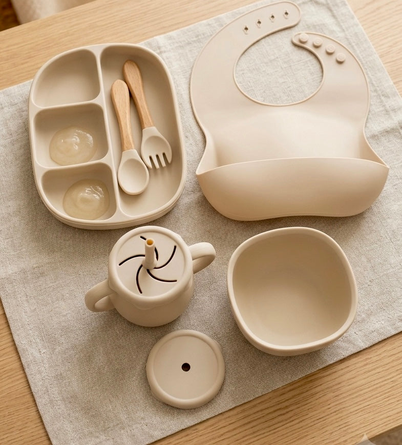 A coordinated baby feeding set arranged on a light fabric surface, including a divided tray with two small compartments, a silicone bib, a small cup with lid, a bowl, and utensils with wooden handles.