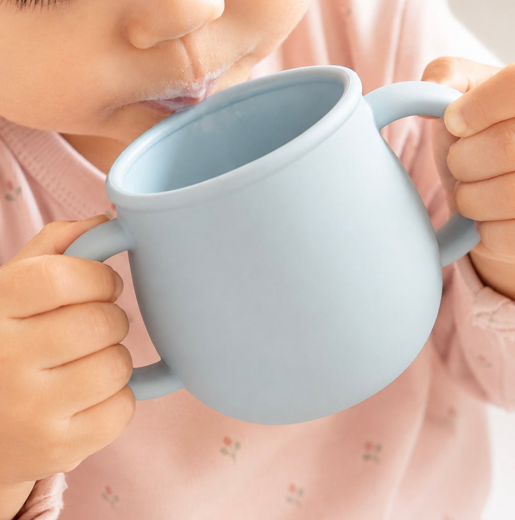 A close-up of a child drinking from a light blue silicone cup with two handles, wearing a pink floral long-sleeve top.