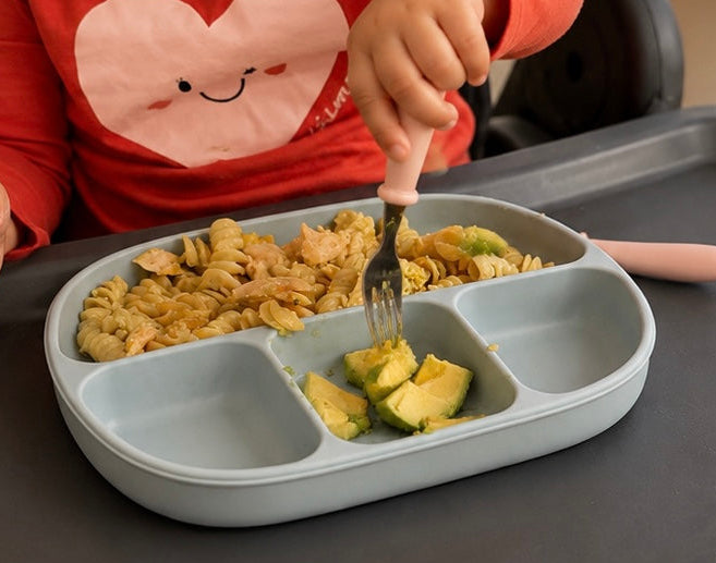 A close-up of a child wearing a red long-sleeve shirt with a pink heart graphic, sitting in a high chair and using a pink-handled fork to eat from a light blue divided meal tray on a dark tray table.