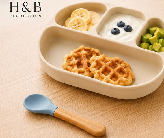 A beige divided children's plate with waffles, yogurt, blueberries, and avocado, shown on a wooden table with a blue-handled spoon. 