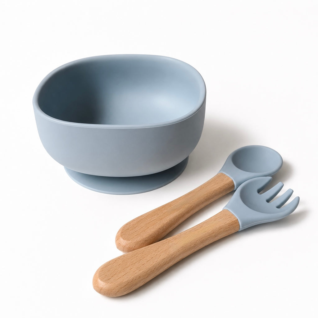 A light blue silicone baby bowl with a suction base, accompanied by a matching silicone spoon and fork with natural wood handles, on a clean white background.