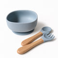 A light blue silicone baby bowl with a suction base, accompanied by a matching silicone spoon and fork with natural wood handles, on a clean white background.