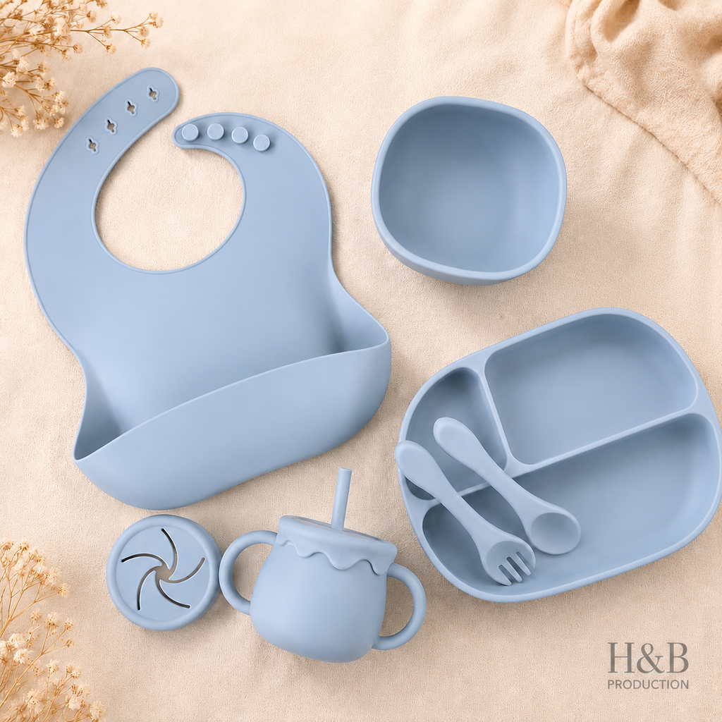 A light blue silicone baby feeding set arranged on a beige fabric background, including a bib, bowl, divided plate, sippy cup, lid, and utensils. The H&B Production logo appears in the bottom right corner.