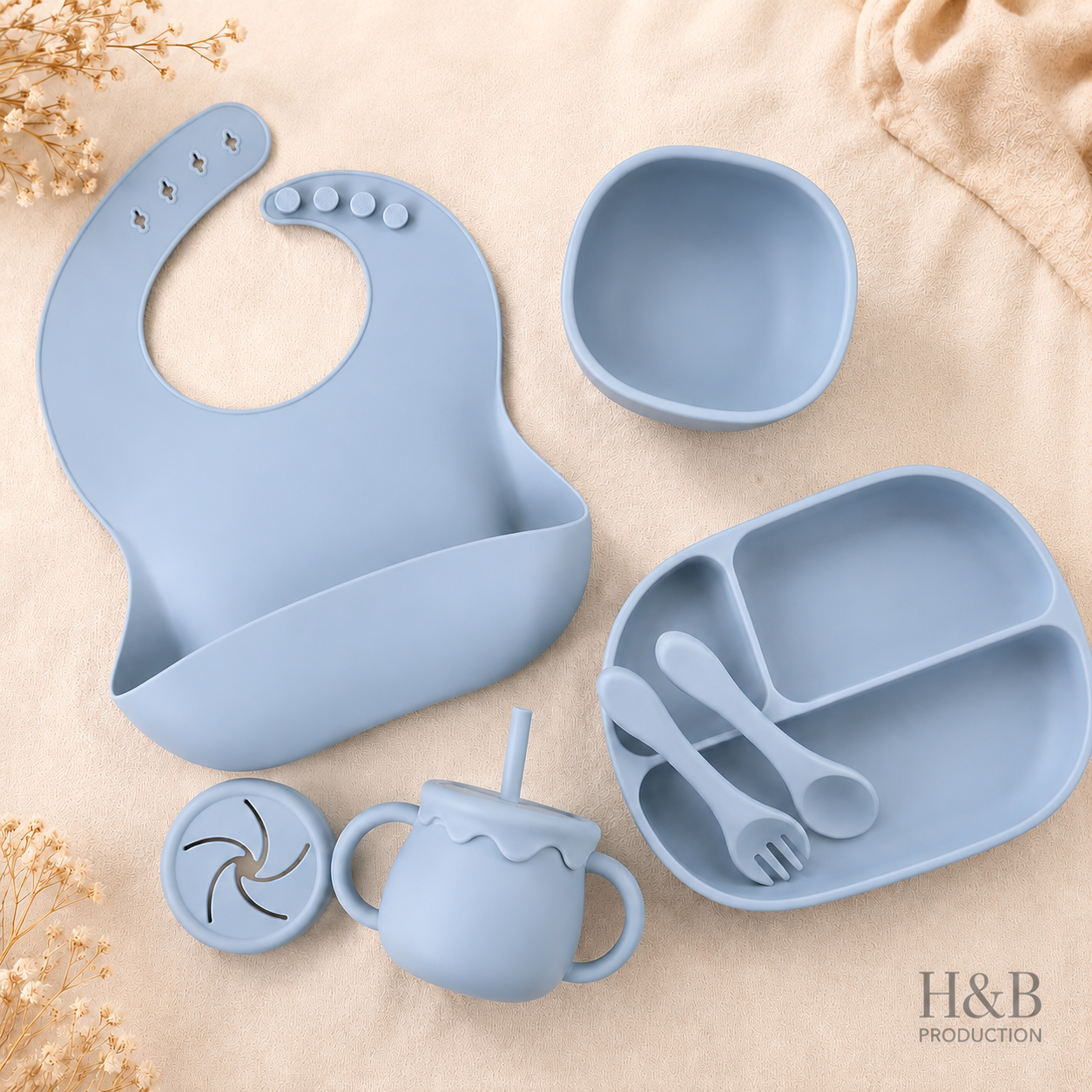A light blue silicone baby feeding set arranged on a beige fabric background, including a bib, bowl, divided plate, sippy cup, lid, and utensils. The H&B Production logo appears in the bottom right corner.