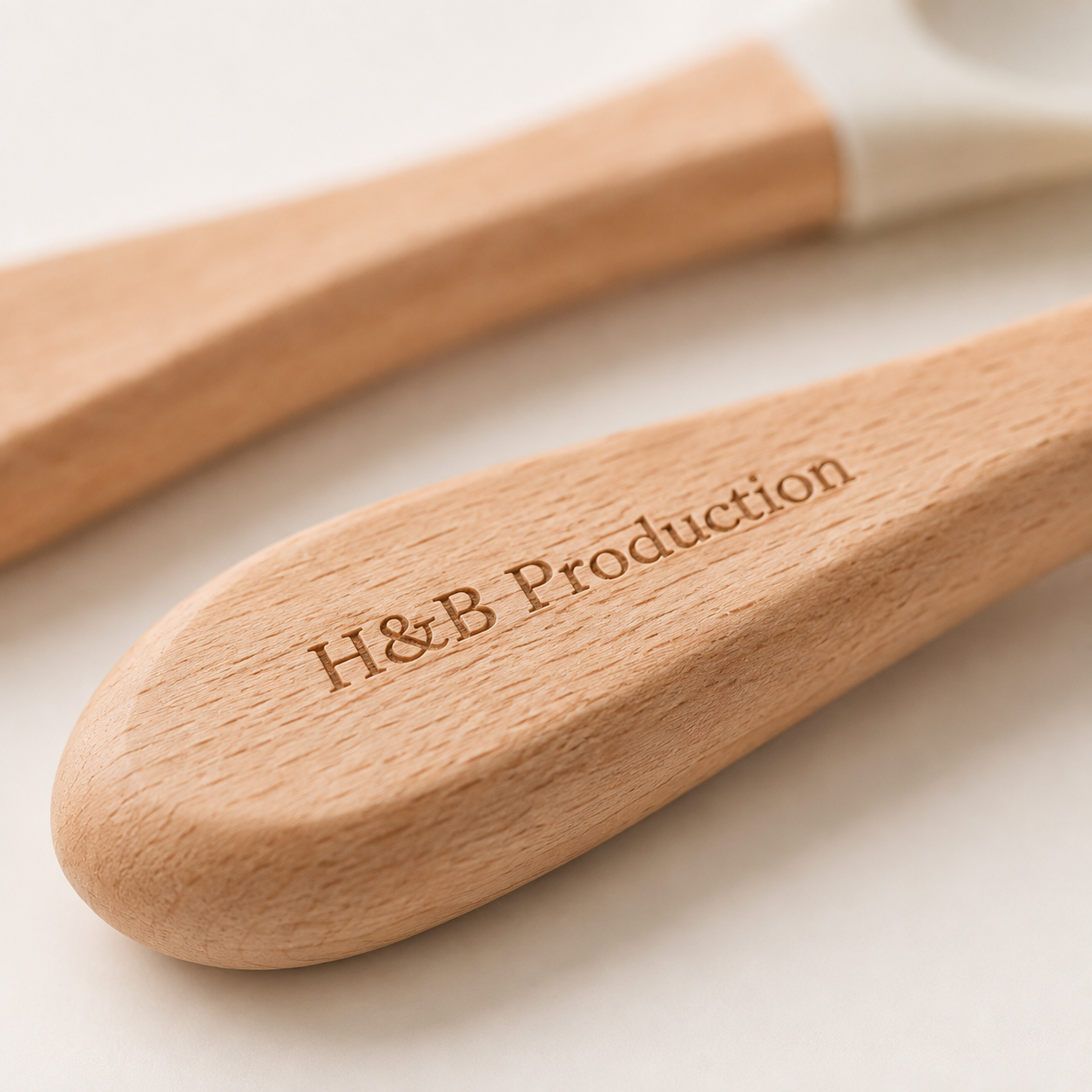 Close-up of two wooden kitchen utensils with white silicone tips, featuring the engraved brand name 'H&B Production' on the foreground utensil.