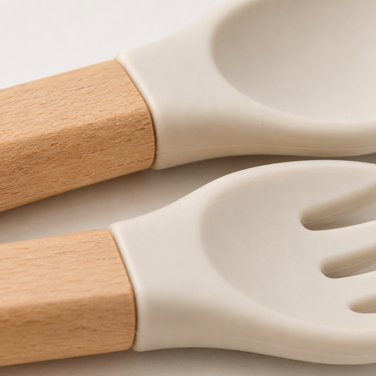 Close-up of a silicone utensil set with light natural wood handles and off-white silicone heads, including a spoon and a fork, on a light neutral background.