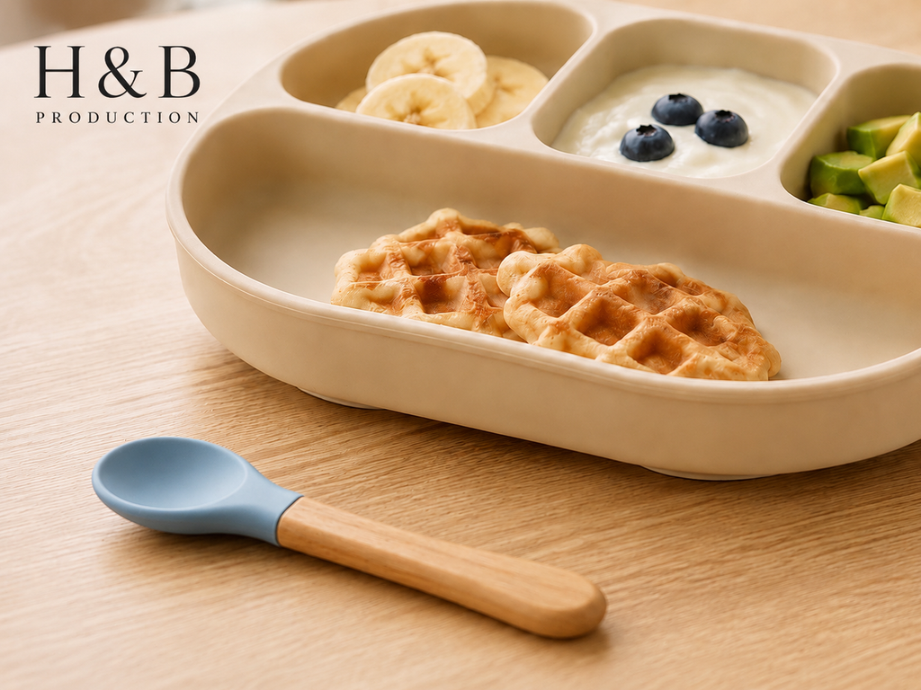 A beige compartment baby meal tray with waffles, yogurt with blueberries, banana slices, and avocado, shown on a wooden table with a blue and wooden spoon. H&B PRODUCTION logo is visible in the top left.