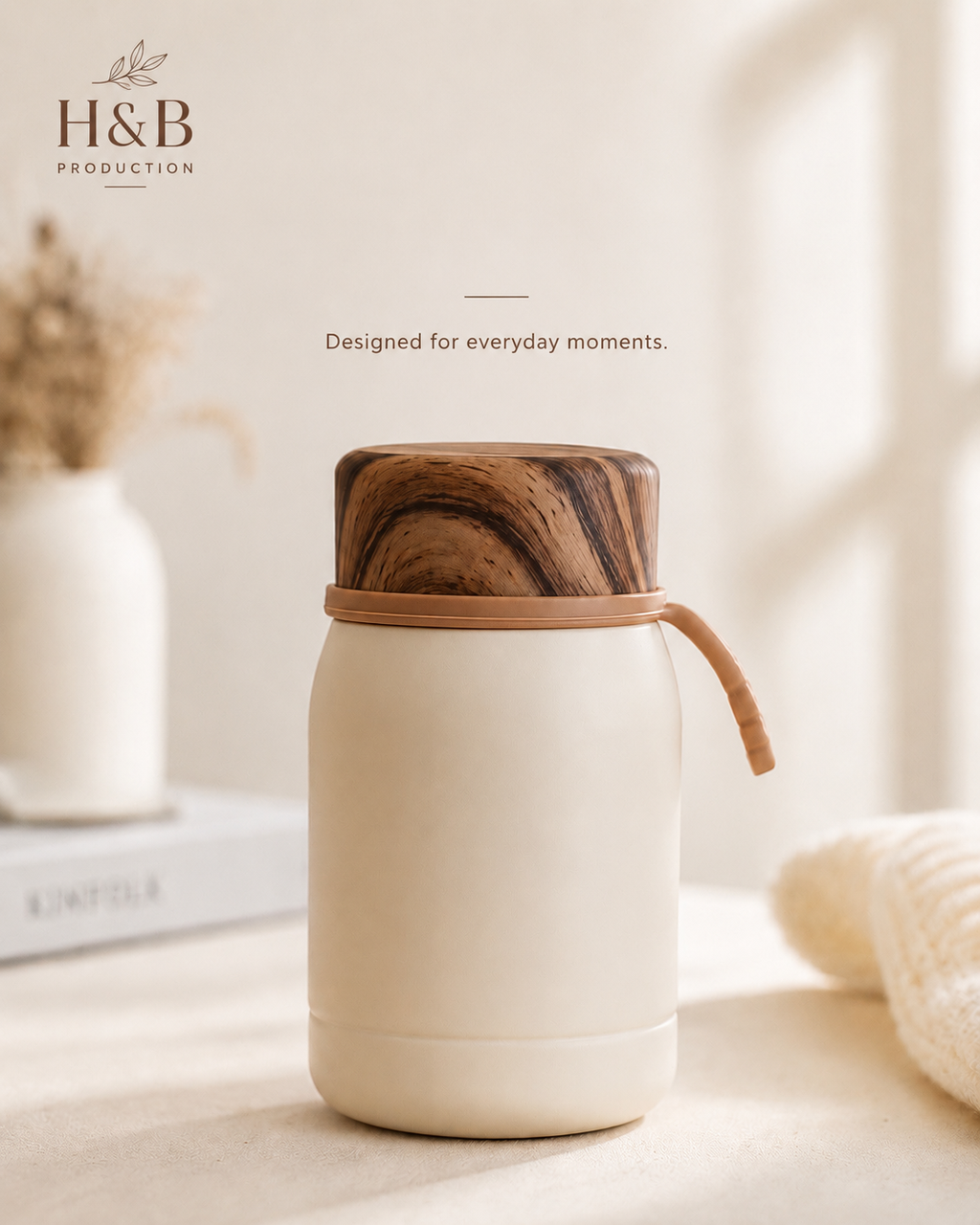 An H&B Production insulated bottle with a cream-colored body and a dark brown wood-grain lid, shown on a tabletop with a knit blanket and a vase in the background.